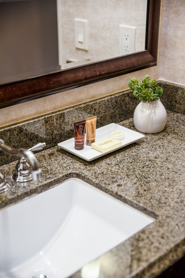 A bathroom sink with a granite countertop, a mirror, and toiletries including a tube, bar of soap, and a small potted plant. - Ayres Suites Diamond Bar (Diamond Bar)