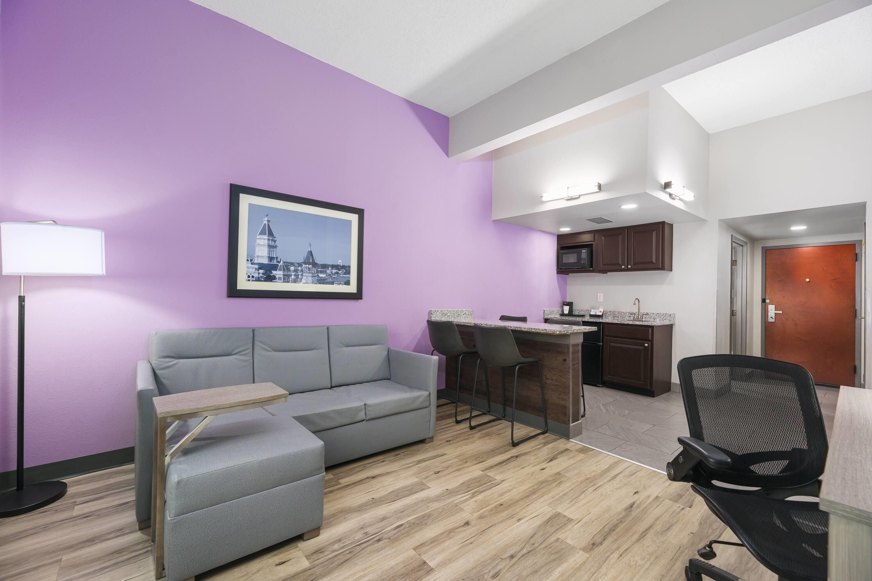 Un salon moderne avec un canapé gris, une petite table, un fauteuil de bureau noir, un coin cuisine avec un bar et un tableau encadré au mur.