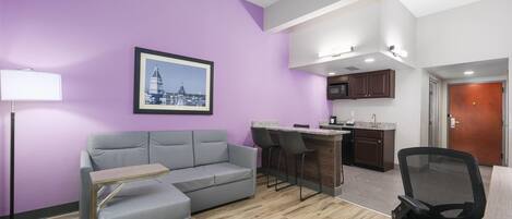 Un salon moderne avec un canapé gris, une petite table, un fauteuil de bureau noir, un coin cuisine avec un bar et un tableau encadré au mur.