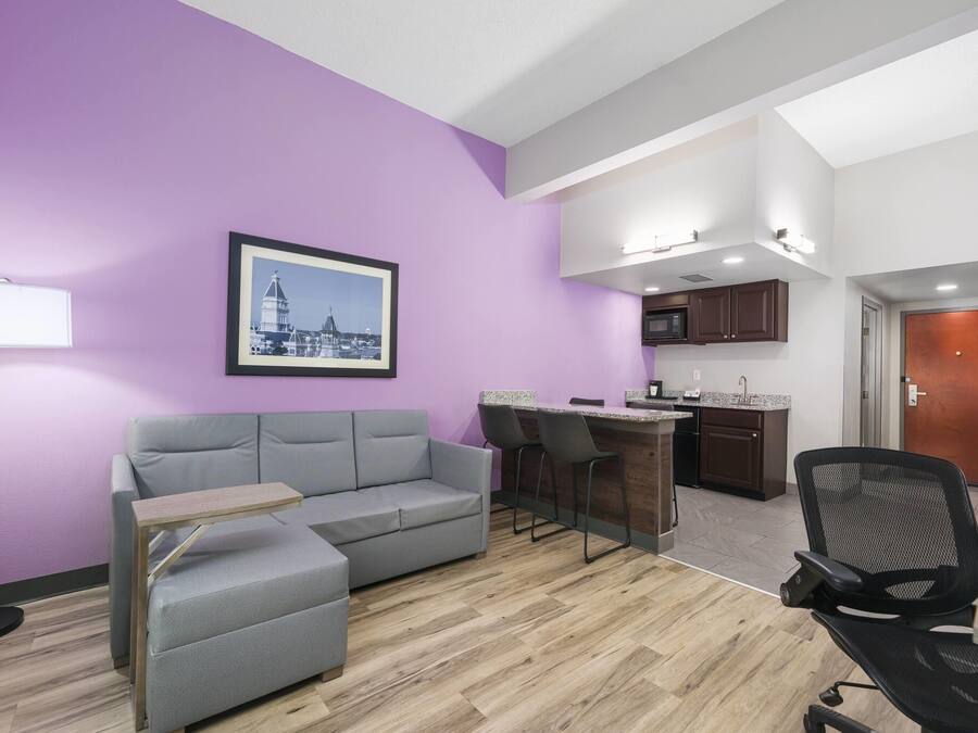 Ein modernes Wohnzimmer mit grauem Sofa, einem kleinen Tisch, einem schwarzen Bürostuhl, einer Küchenzeile mit Bar und einem gerahmten Bild an der Wand.