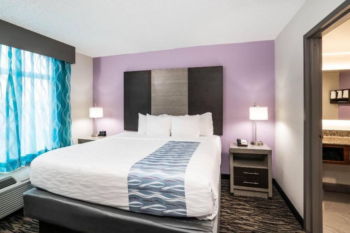 A hotel room with a bed, bedside lamps, a nightstand, a window with curtains, and a bathroom visible through an open door.