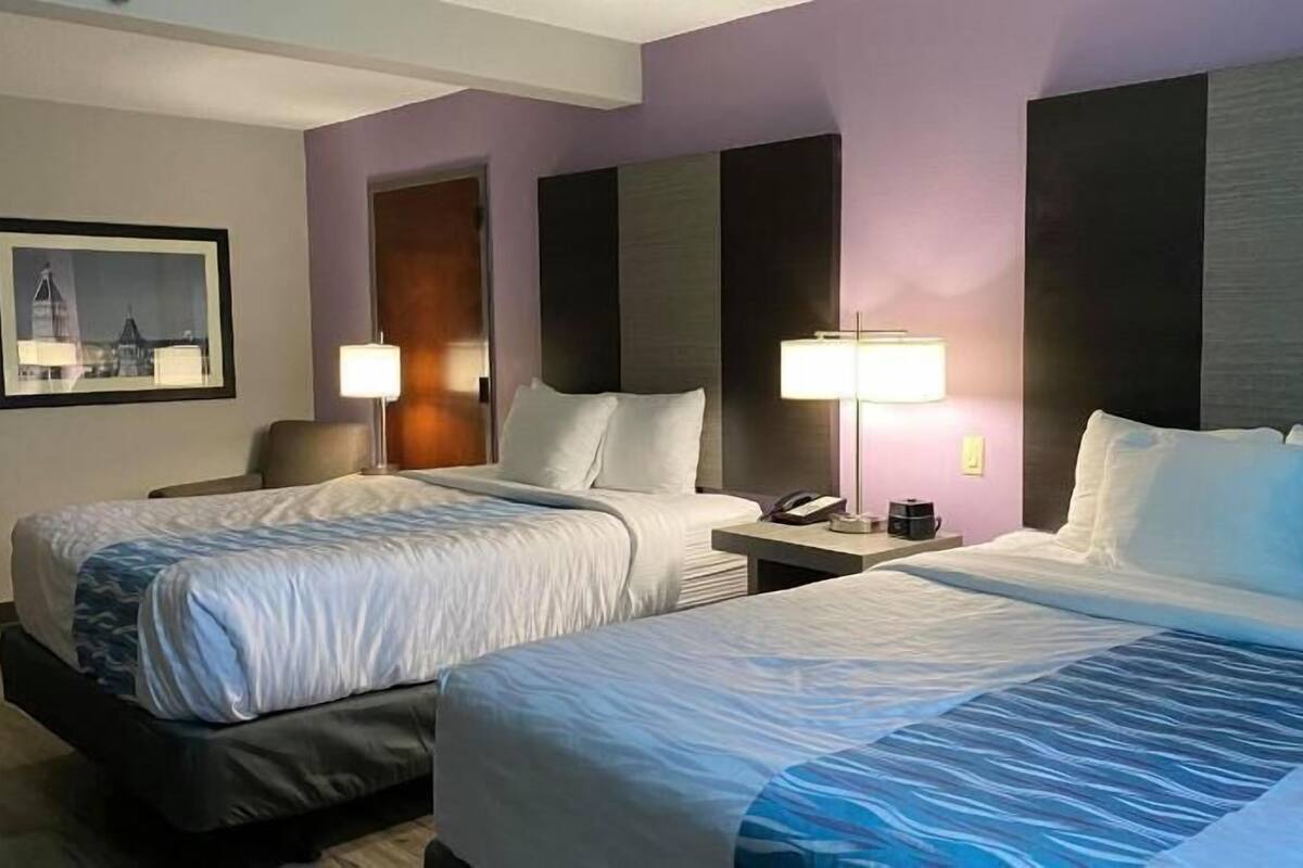 A hotel room with two beds, a nightstand with a lamp, a chair, and a framed picture on the wall.