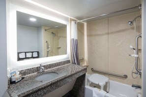 Un baño con lavabo de encimera de granito, espejo, ducha con barra de apoyo y inodoro blanco.