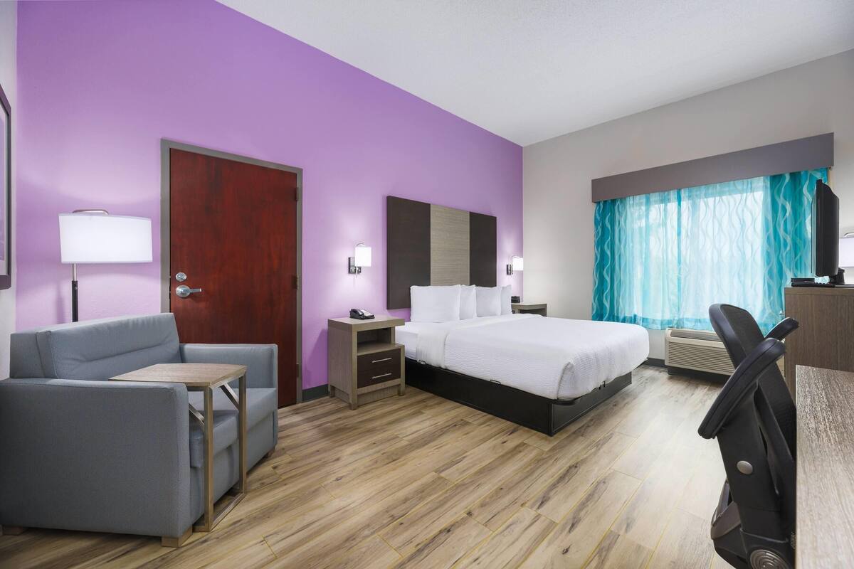 Habitación de hotel con cama, sofá, escritorio, silla, mesita de noche, lámpara, espejo, ventana con cortinas y equipo de aire acondicionado.
