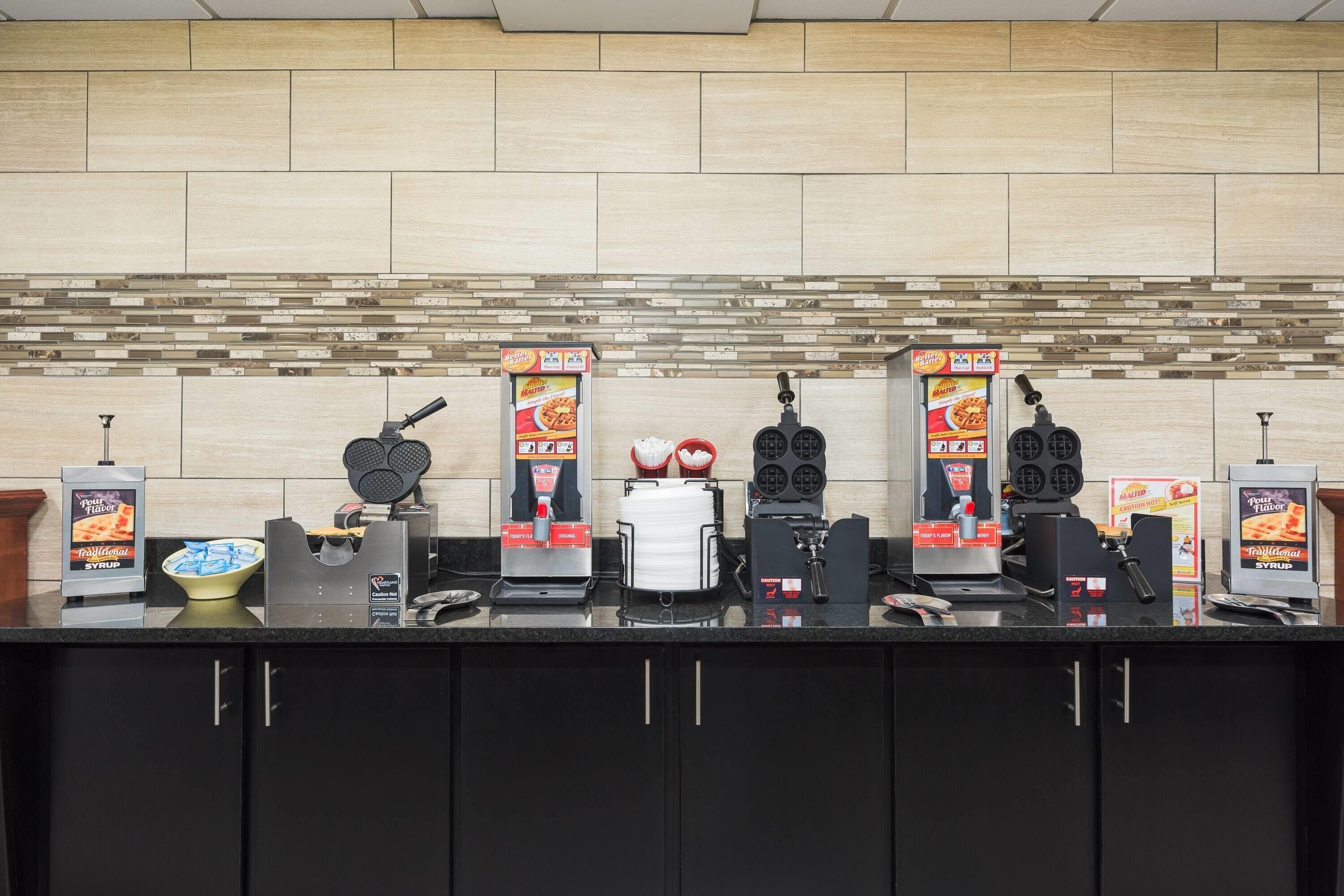 A counter with multiple coffee machines and a waffle maker, a bowl of chips, and a sign for 'Pour Your Flavor Syrup'.