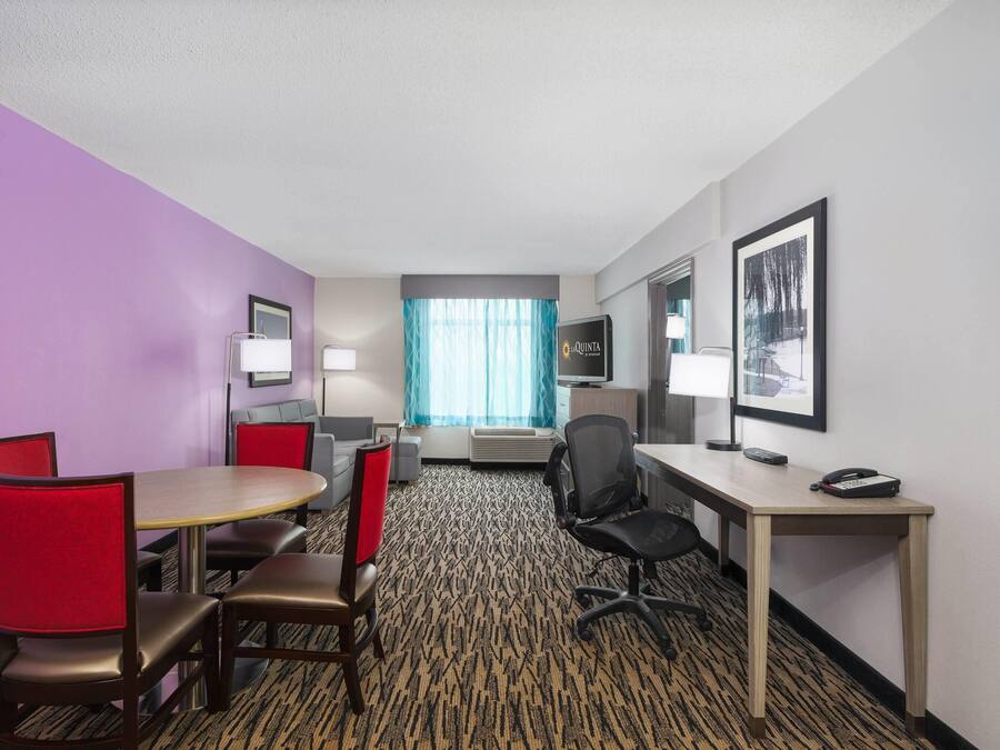A hotel room with a purple wall, a wooden desk, a red chair, a black office chair, a round table, a TV, and a framed picture on the wall.