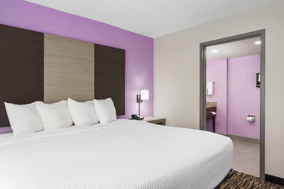 Habitación de hotel con una cama grande, una mesita de noche con lámpara, baño visible a través de una puerta abierta y una pared de acento morada.