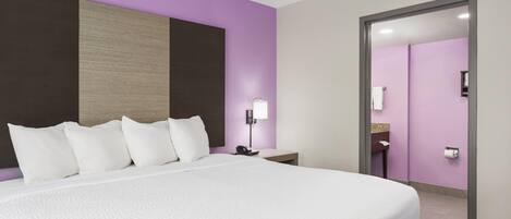 A hotel room with a large bed, a nightstand with a lamp, a bathroom visible through an open door, and a purple accent wall.