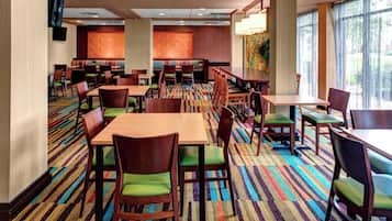 A hotel dining area with multiple tables and chairs, a patterned carpet, and large windows allowing natural light.