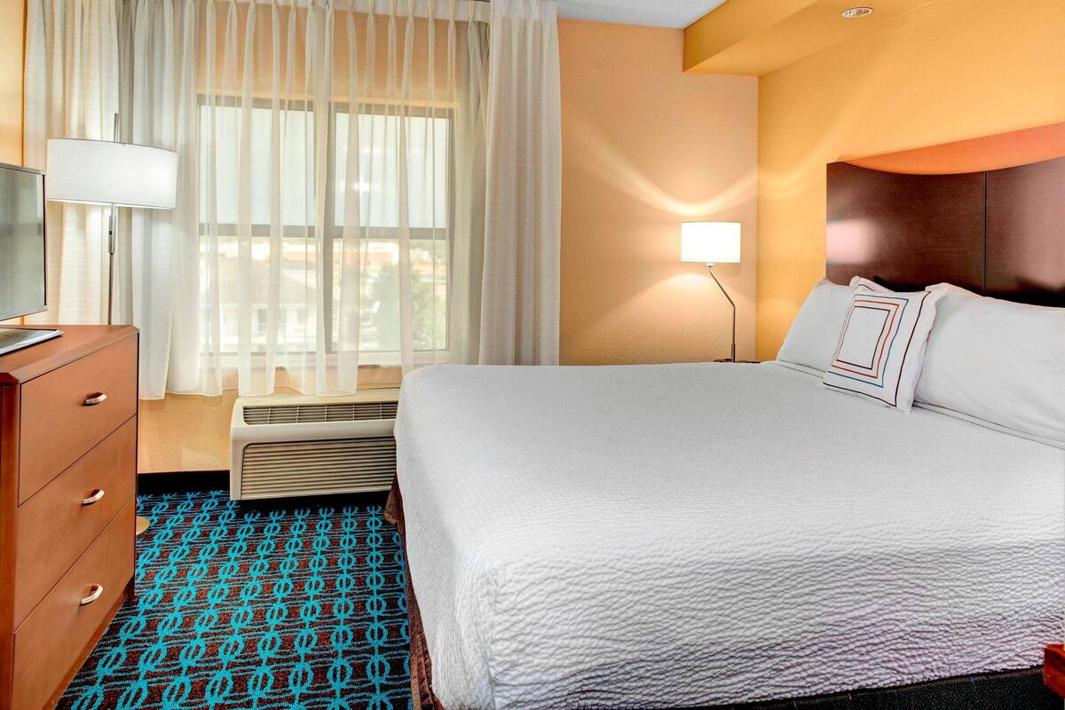 A hotel room with a bed, a dresser, a window with curtains, a wall-mounted lamp, and a patterned carpet.