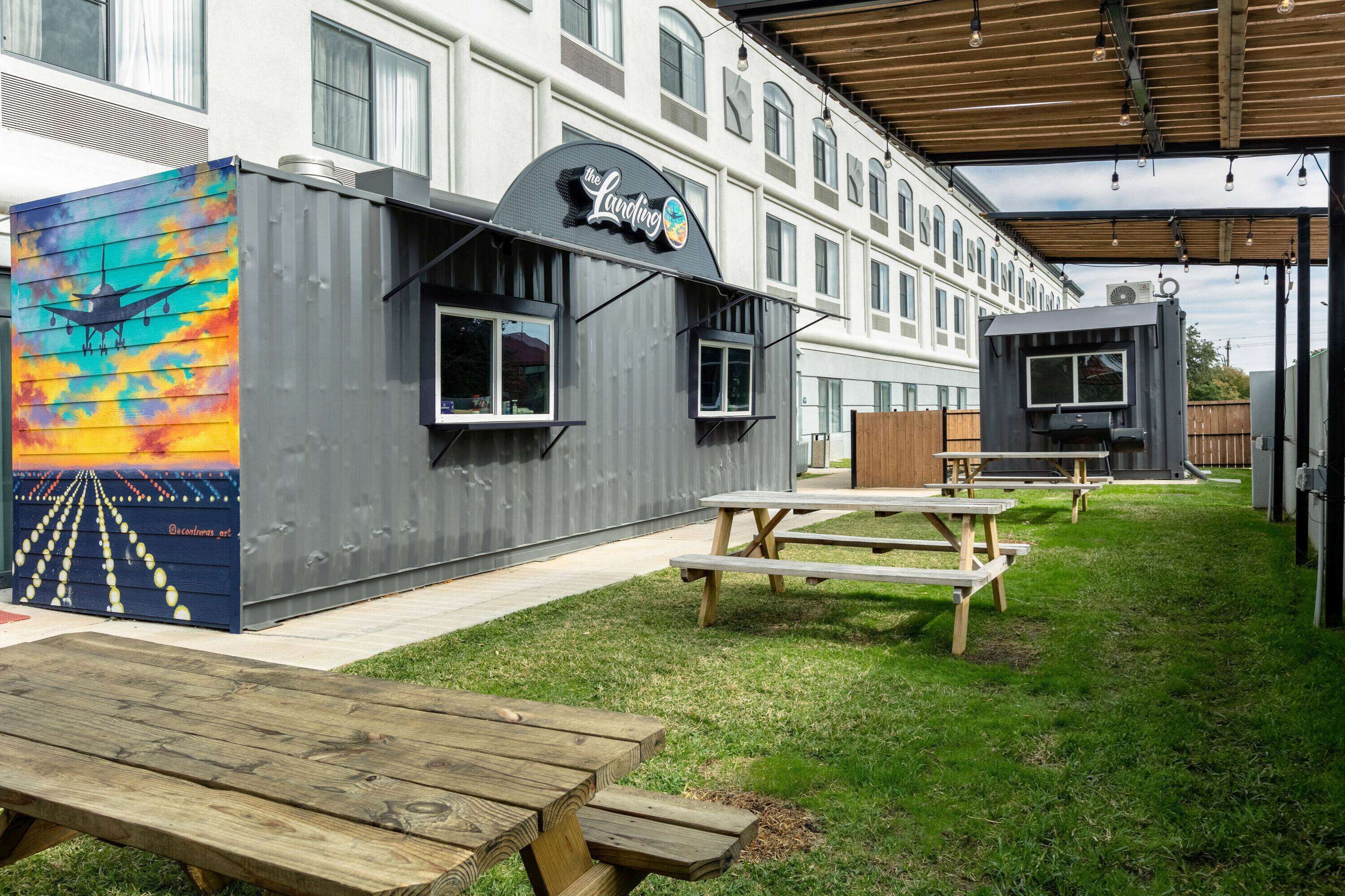 A building with a restaurant sign, outdoor wooden picnic tables, and a colorful mural on a container.