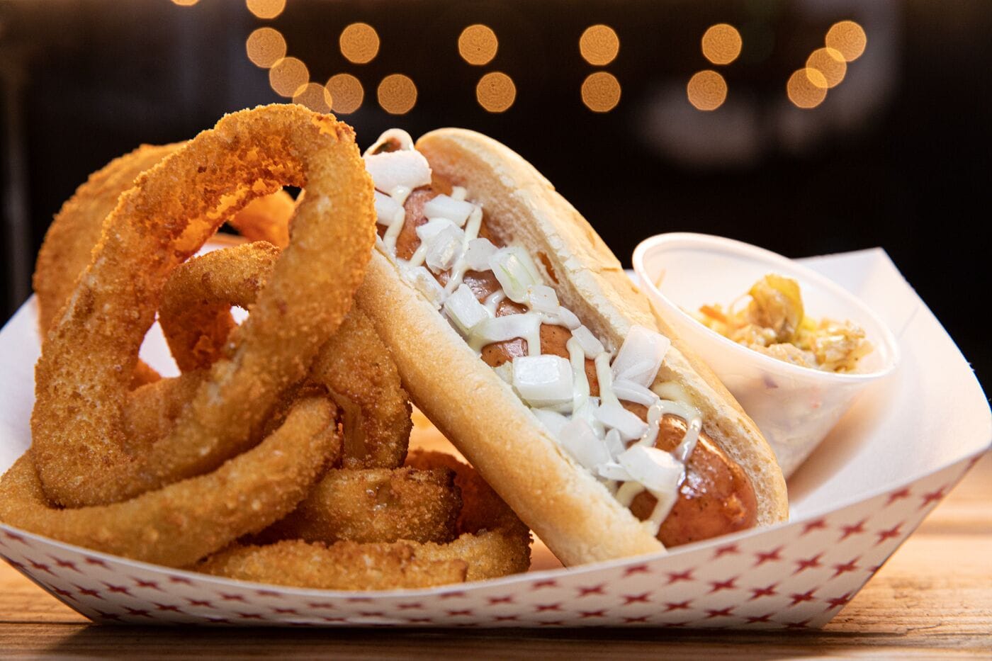 A hot dog with onions and a bun, accompanied by onion rings and a side of coleslaw in a paper tray.
