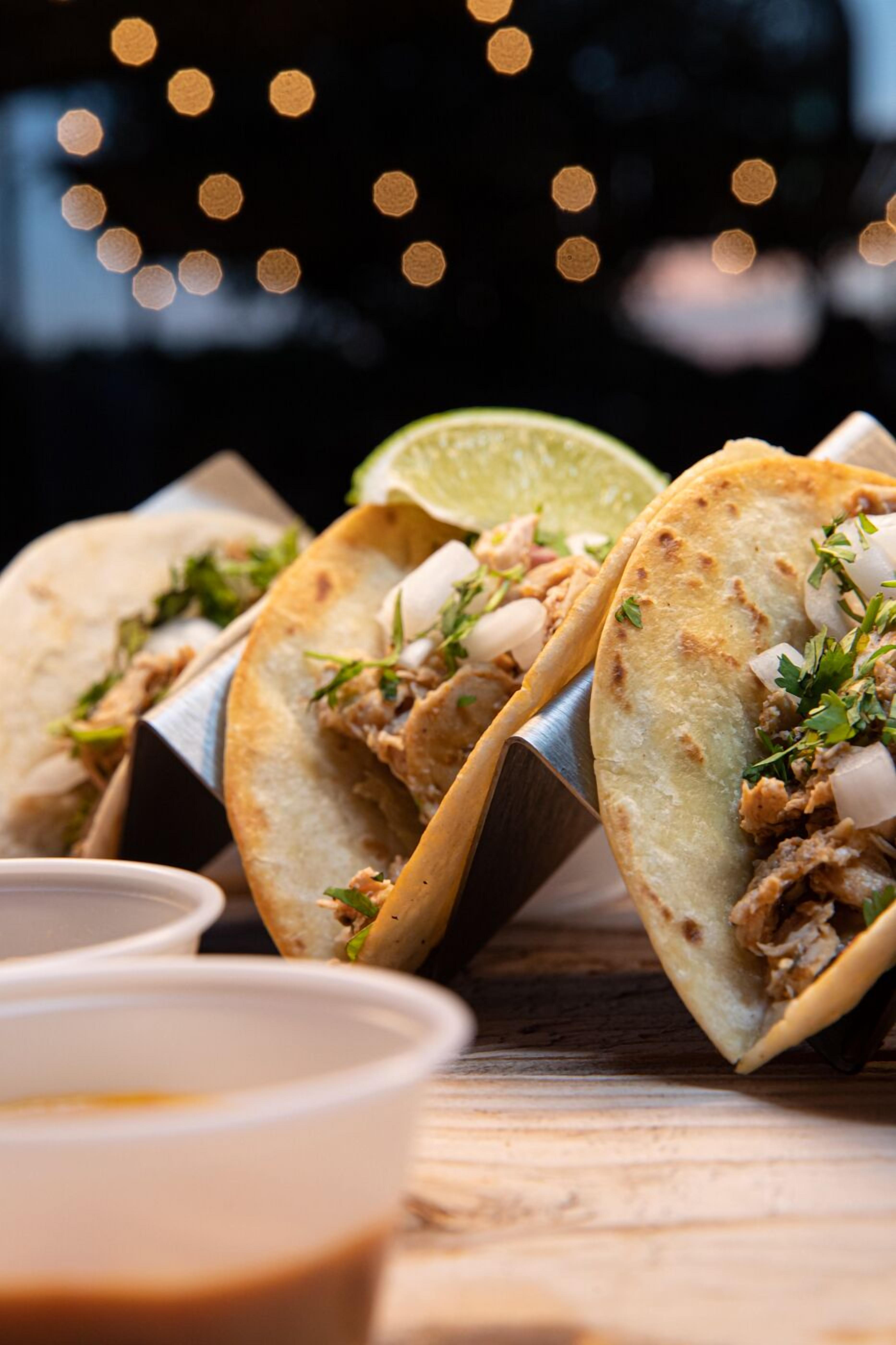 Three tacos with meat, onions, and cilantro, accompanied by a lime wedge and a small bowl of sauce.