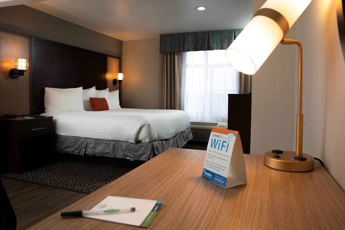 Ein Hotelzimmer mit Bett, Nachttischlampen, einem Schreibtisch mit WLAN-Symbol und einem Fenster mit Vorhängen.