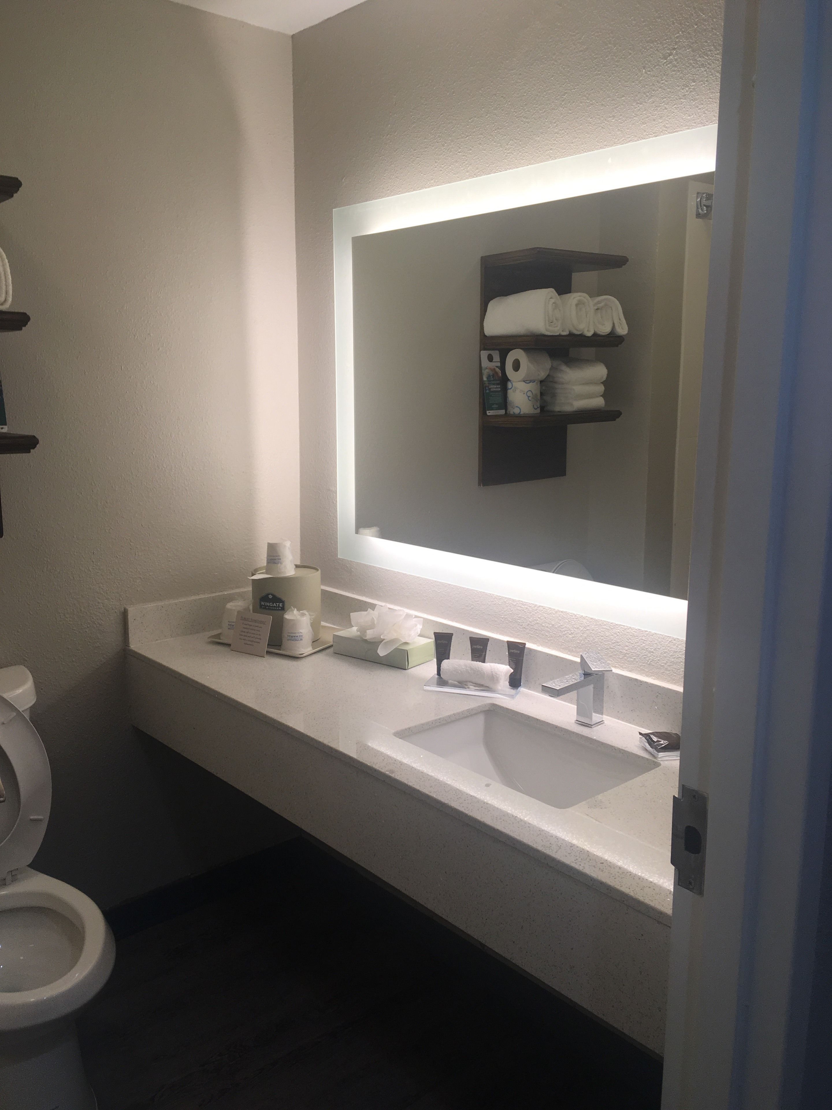 Un baño con lavamanos blanco, un espejo y un estante con toallas y artículos de aseo.