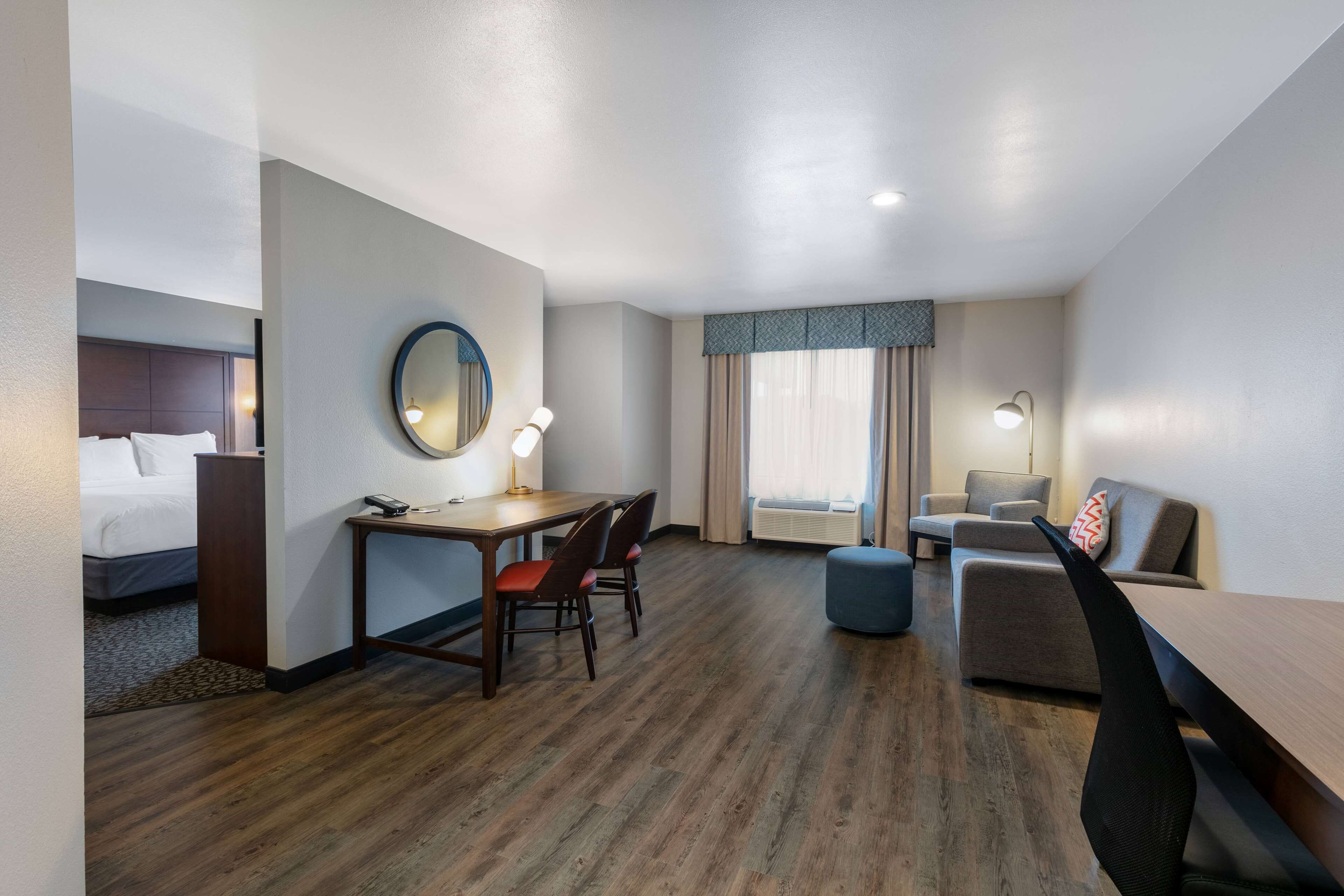 A hotel room with a bed, a desk with a chair, a round mirror, a small table, a sofa, and a window with curtains.