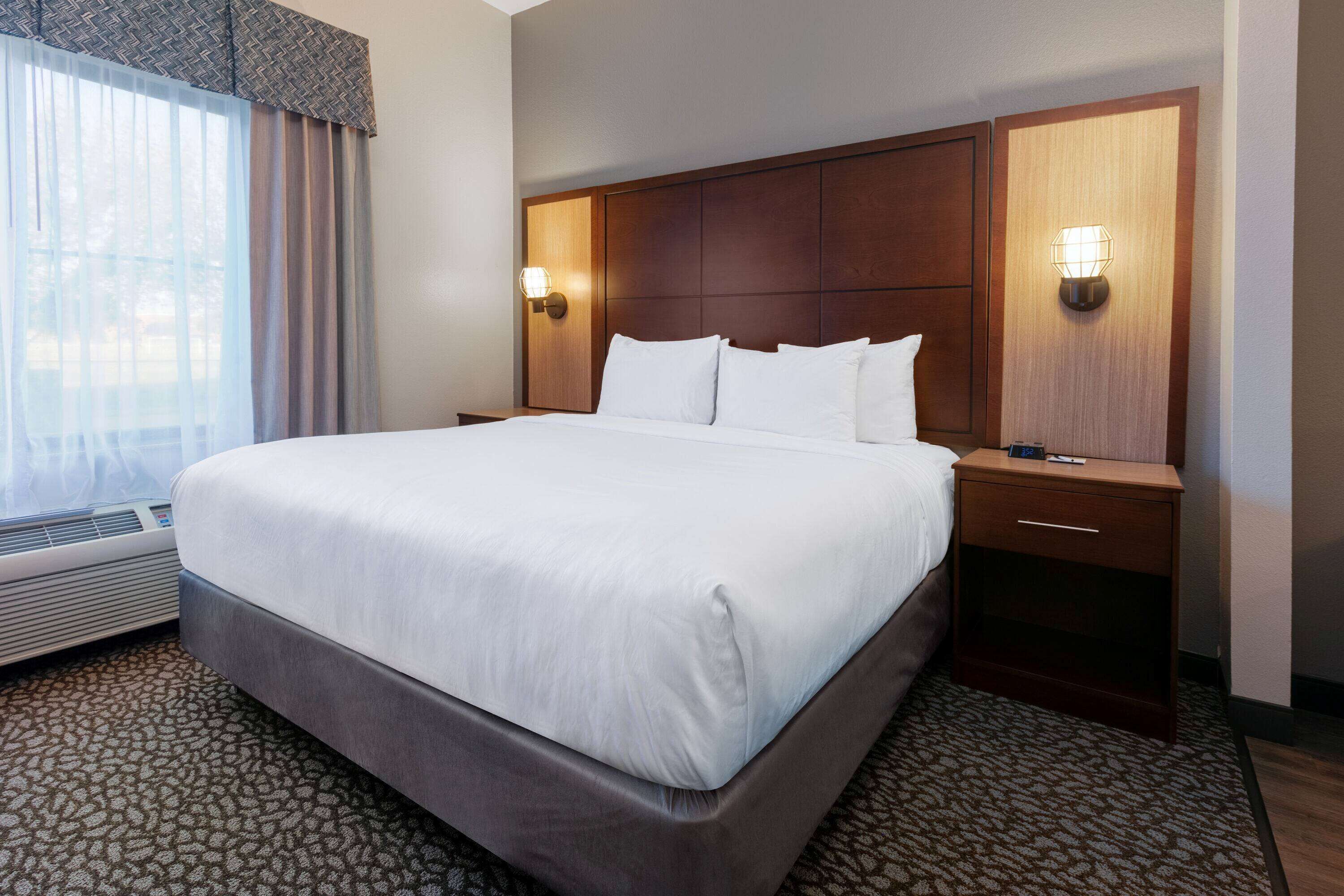 Una cama grande con cabecera de madera, dos mesitas de noche con lámparas, una ventana con cortinas traslúcidas y una alfombra estampada.