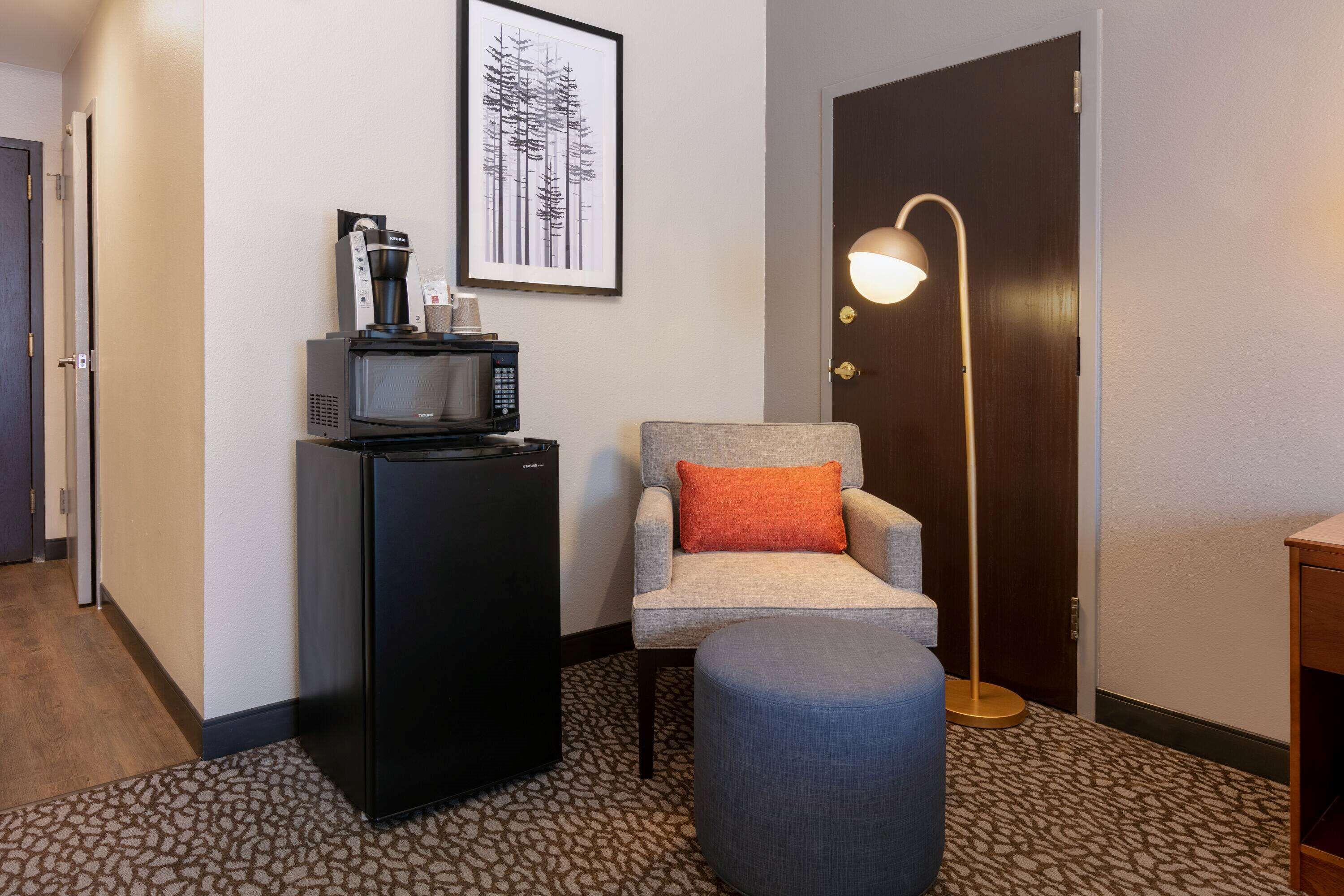Een hotelkamer met kitchenette, een grijze fauteuil met oranje kussen, een staande vloerlamp en een kamerbreed tapijt met patroon.