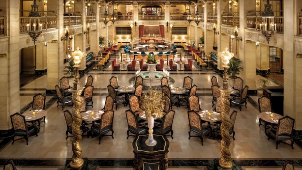 A grand hotel lobby with ornate chandeliers, a central bar area, and multiple dining tables with chairs. - The Louie, A Davenport Hotel, Autograph Collection® (Spokane)