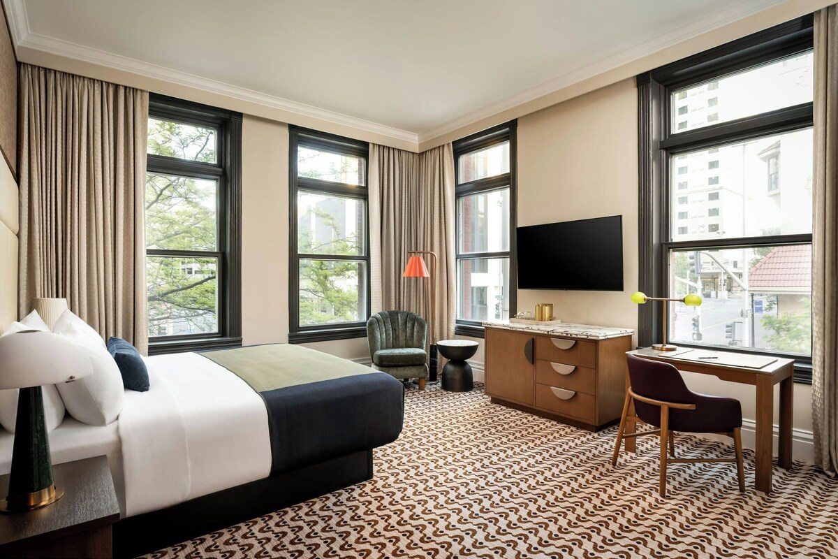 A hotel room with a bed, a desk, a chair, a TV, and large windows.