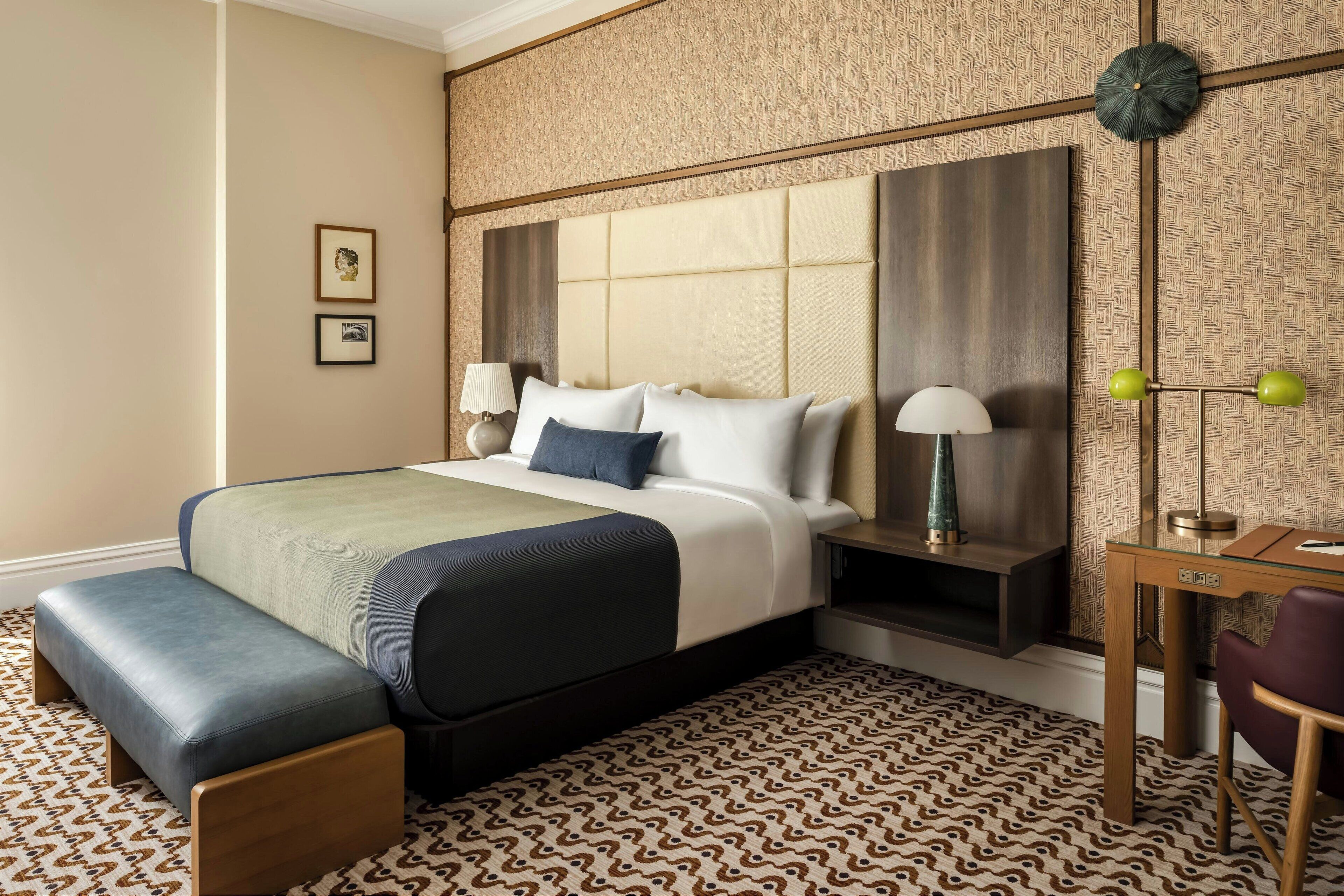 Habitación de hotel con una cama grande, un banco, un escritorio y una silla. Hay dos lámparas en las mesitas de noche y un reloj en la pared.