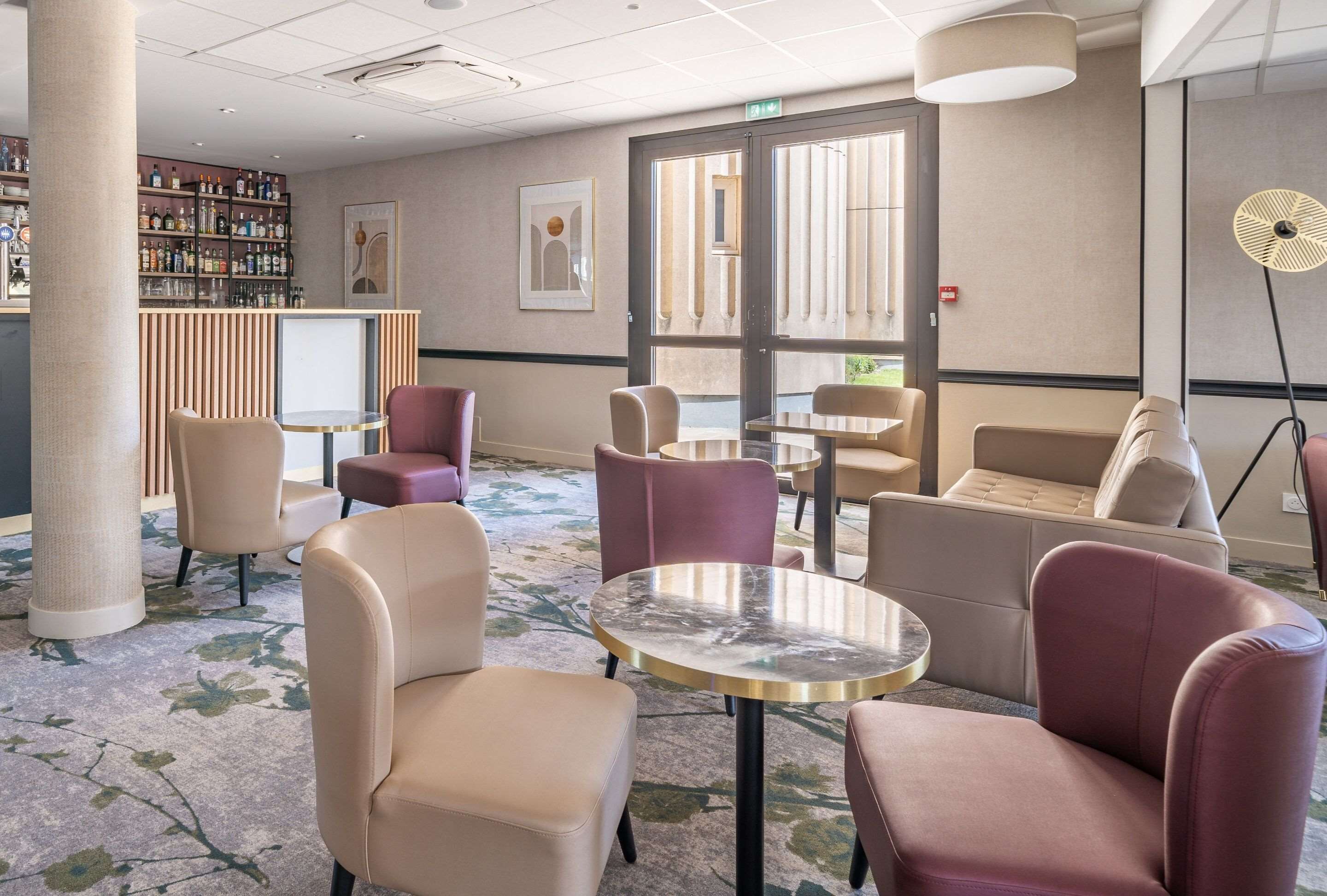 A hotel lobby with seating areas, a bar with a variety of bottles, and a large window providing natural light.