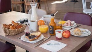 Une table de petit-déjeuner avec des croissants, une bouteille de jus d’orange, un pichet de lait, un bol de céréales et une assiette avec une tranche de pain grillé et de la confiture.