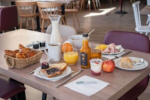 Uma mesa de café da manhã com croissants, uma garrafa de suco de laranja, um jarro de leite, uma tigela de cereal e um prato com uma fatia de torrada e geleia.