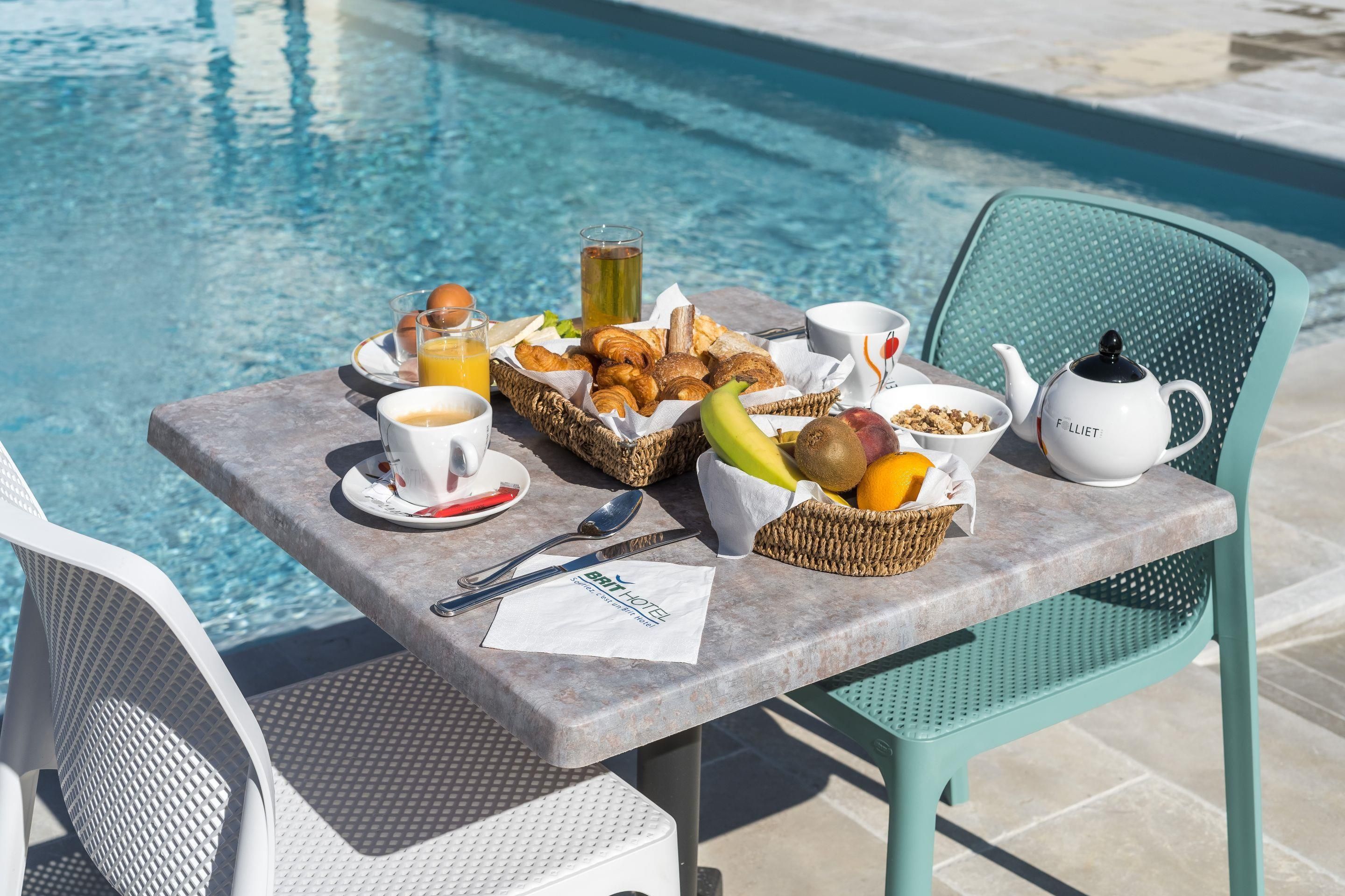A breakfast setup by a poolside with croissants, fruits, and beverages.
