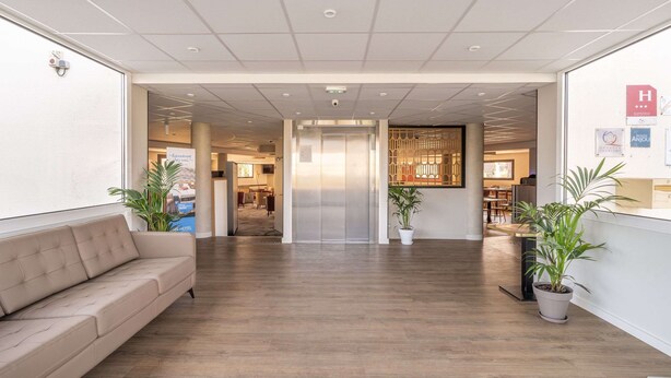 Un hall d’hôtel avec un plancher en bois, un canapé beige, des plantes en pot et un comptoir de réception.