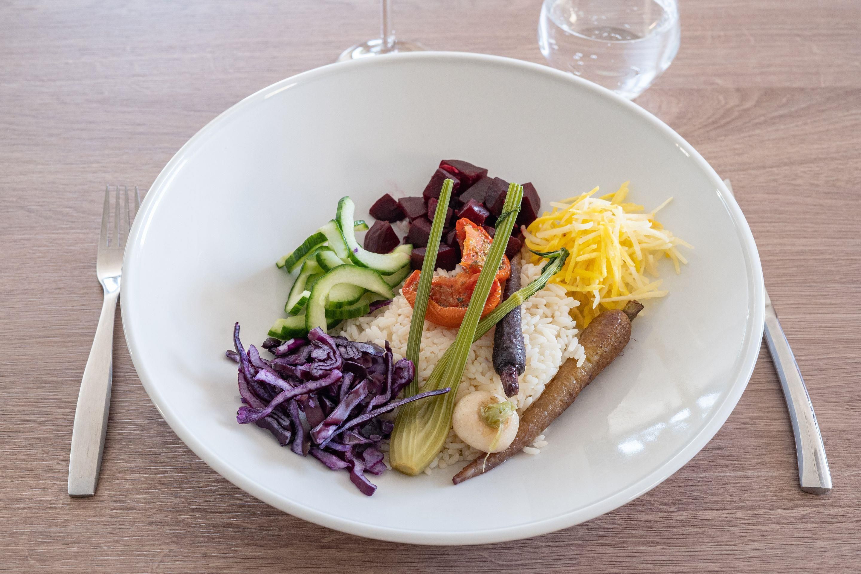 A plate of food with various vegetables and a piece of meat, accompanied by a fork and knife on a wooden table.