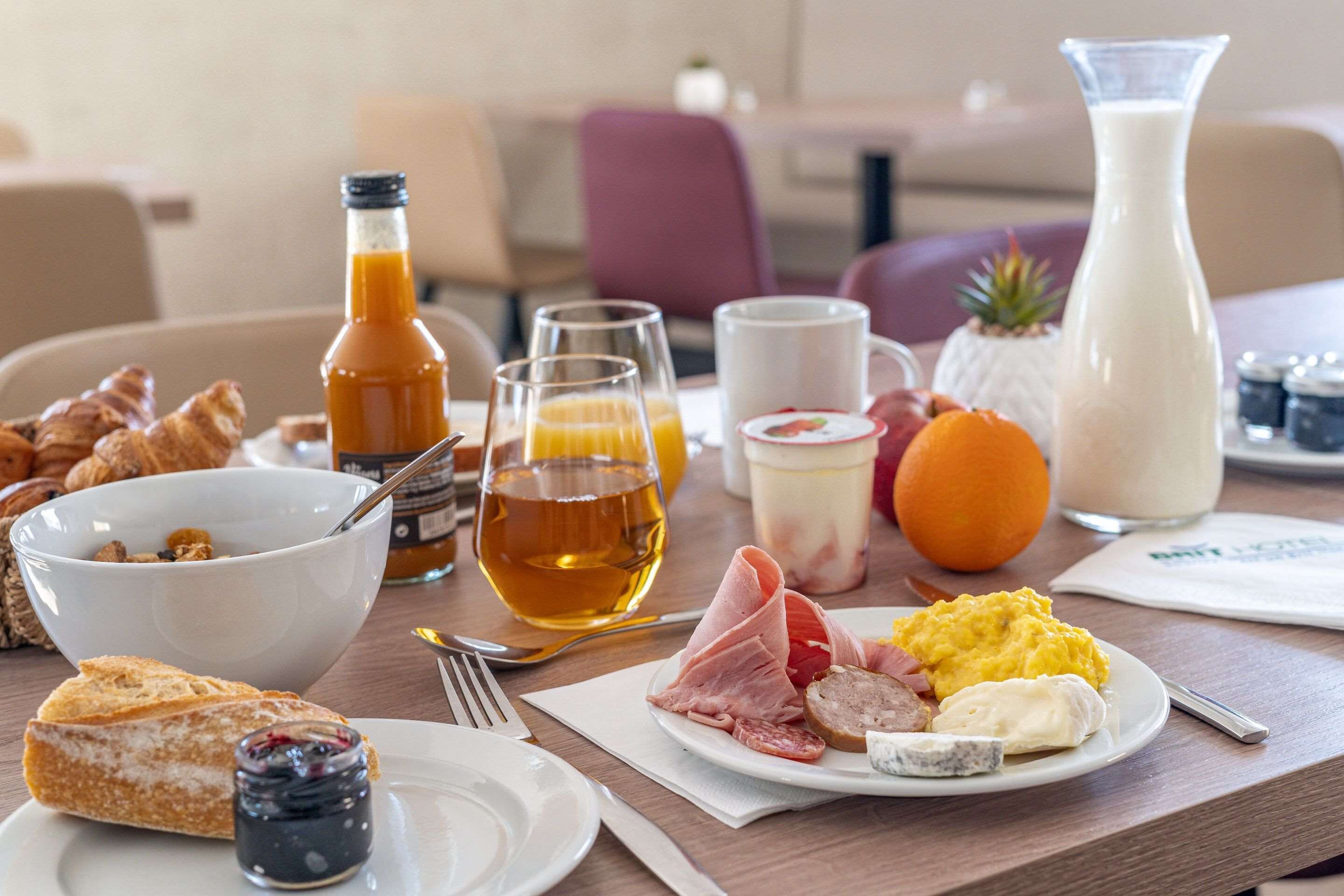 A breakfast table with croissants, a bowl of cereal, a plate of ham, eggs, and a jug of milk.