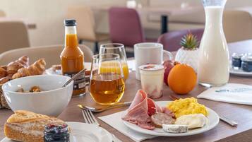 A breakfast table with croissants, a bowl of cereal, a plate of ham, eggs, and a jug of milk.
