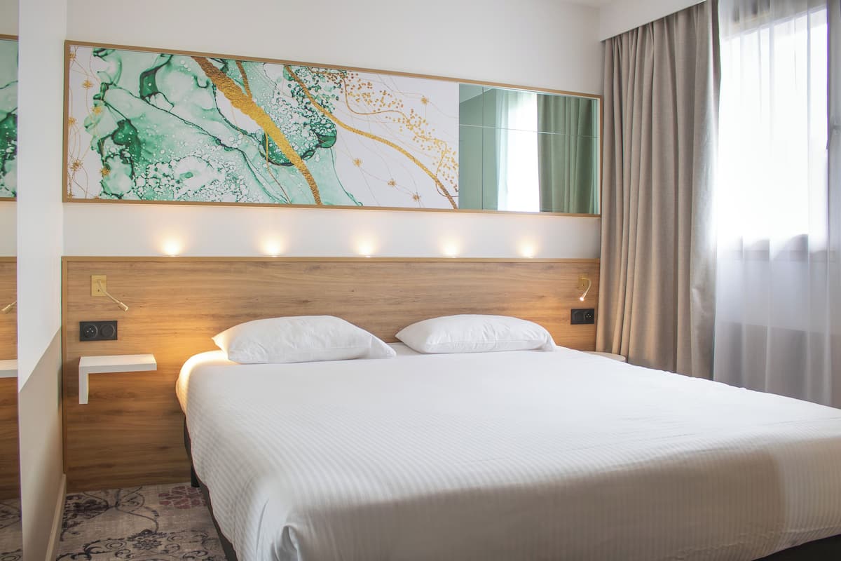 Een hotelkamer met een groot bed, een houten hoofdeinde, een kunstwerk aan de muur en een raam met gordijnen.