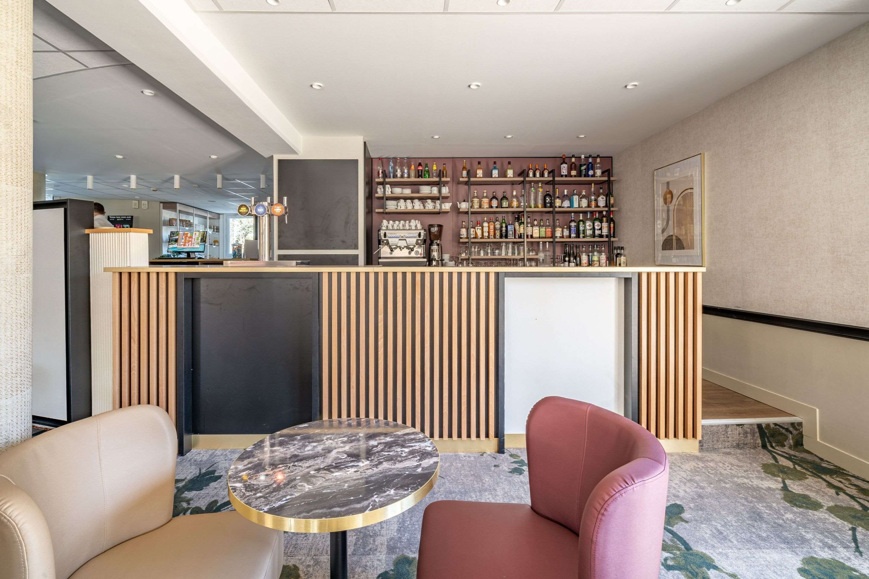 A modern bar area with a wooden counter, shelves of bottles, and a seating area with two chairs and a small round table.