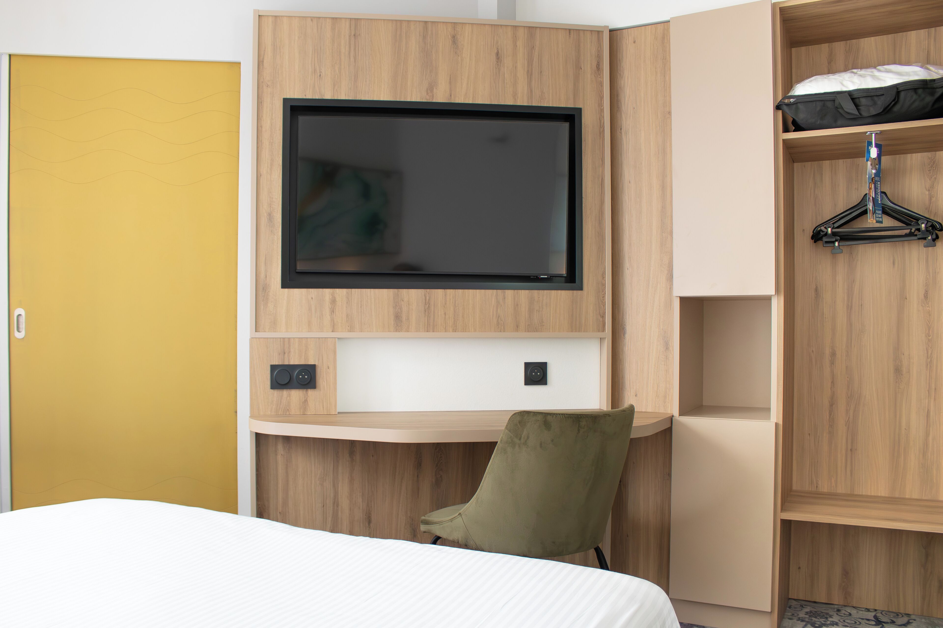 A hotel room with a bed, a TV mounted on the wall, a wooden desk, and a wardrobe.