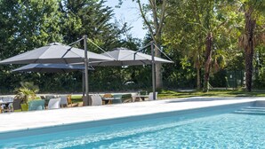 Une piscine agrémentée de deux parasols pour créer de l’ombre, entourée de chaises longues et de tables, avec des arbres en arrière-plan.