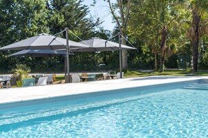 Piscina com duas ombrelhas oferecendo sombra, rodeada por espreguiçadeiras e mesas, com árvores ao fundo.