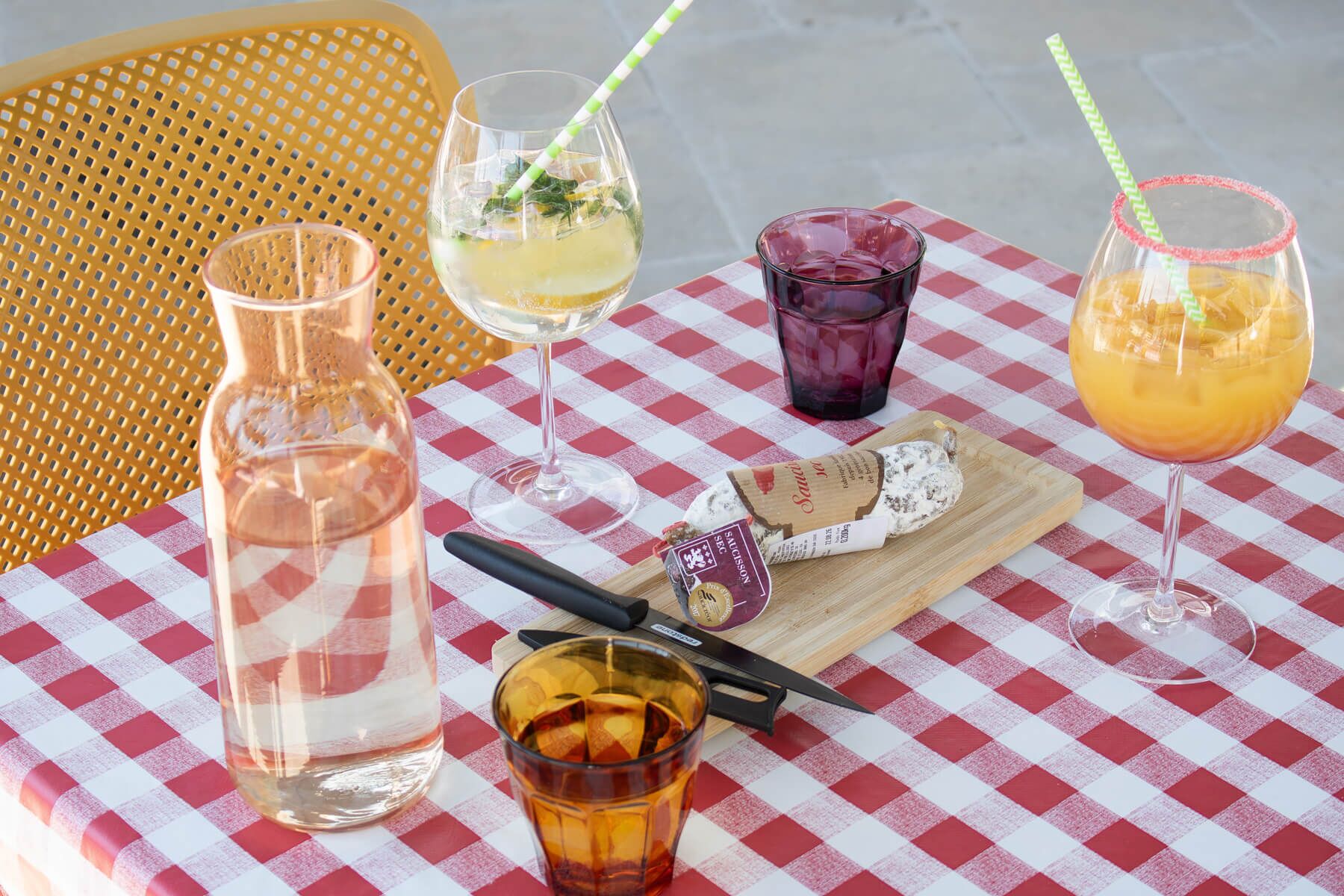 A table with a checkered tablecloth, a bottle of water, a glass of white wine, a glass of red wine, a glass of orange juice, a knife, a cheese board, and a corkscrew.