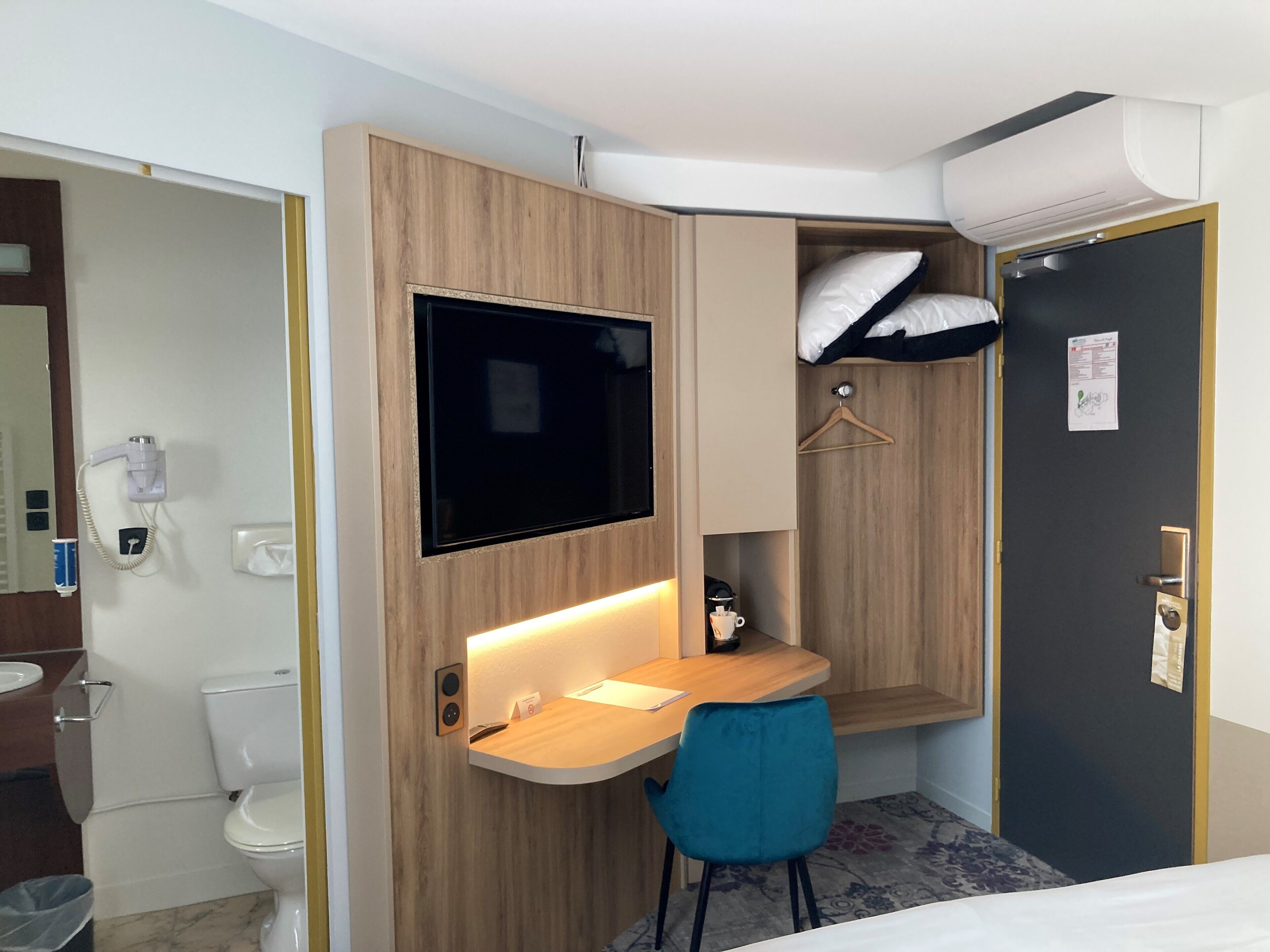 A compact hotel room with a wooden desk, a TV mounted on the wall, a bed, and a small seating area with a chair.