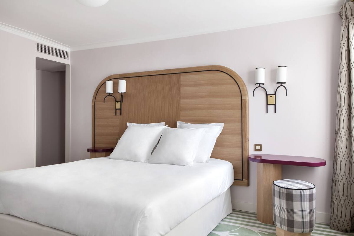 A neatly made bed with white linens, a wooden headboard, a small bedside table with a lamp, and a patterned waste basket.
