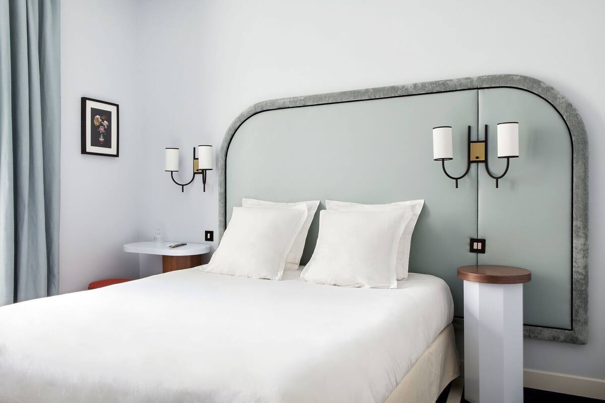 A neatly made bed with white bedding, a round bedside table, a wall-mounted light fixture, and a framed picture on the wall.