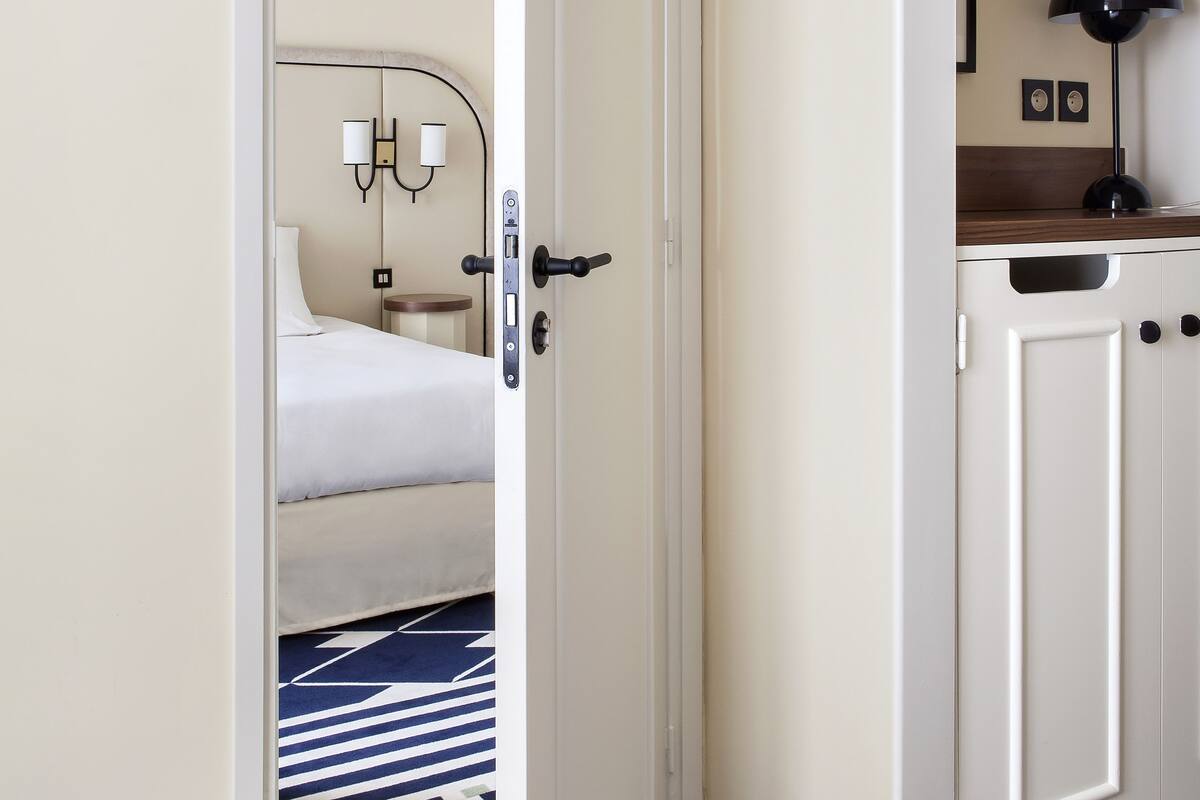 A hotel room with a bed, bedside table, lamp, and a view of the hallway.