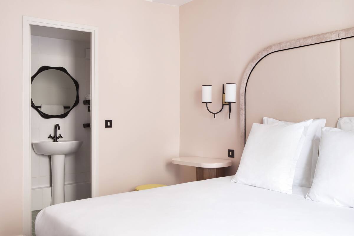 A hotel room with a bed, a bedside table, a small round table, a wall-mounted mirror, and a wall-mounted light fixture.
