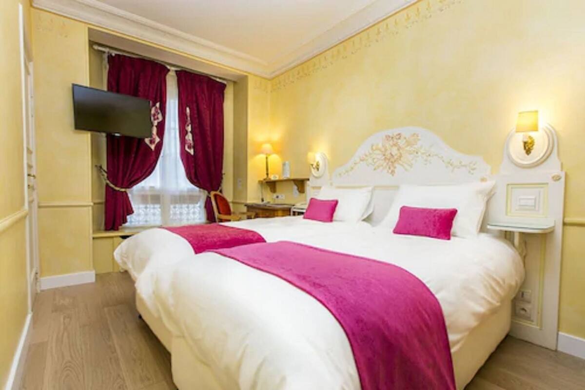 Habitación de hotel con una cama grande, dos mesitas de noche, un televisor montado en la pared y un ventanal con cortinas rojas.