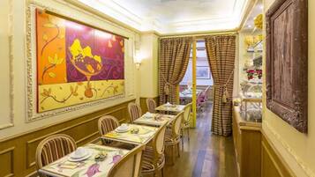 Une salle à manger avec des tables dressées, un grand tableau floral accroché au mur et, à travers les rideaux, une vue sur l’extérieur.
