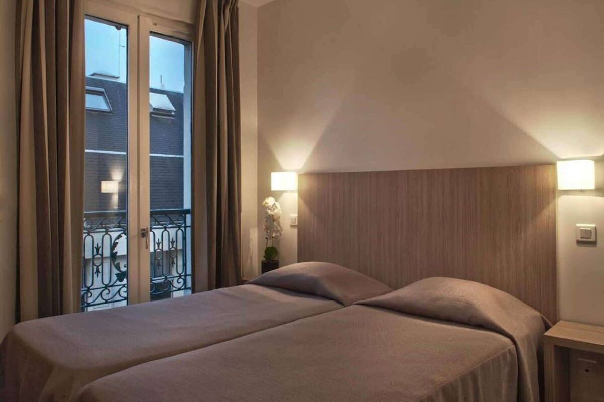 A hotel room with a bed, a nightstand, a wall-mounted lamp, and a window with curtains.