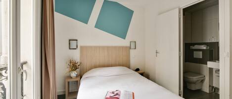 A hotel room with a bed, a nightstand, a wall-mounted shelf with a vase, and a bathroom visible through an open door.