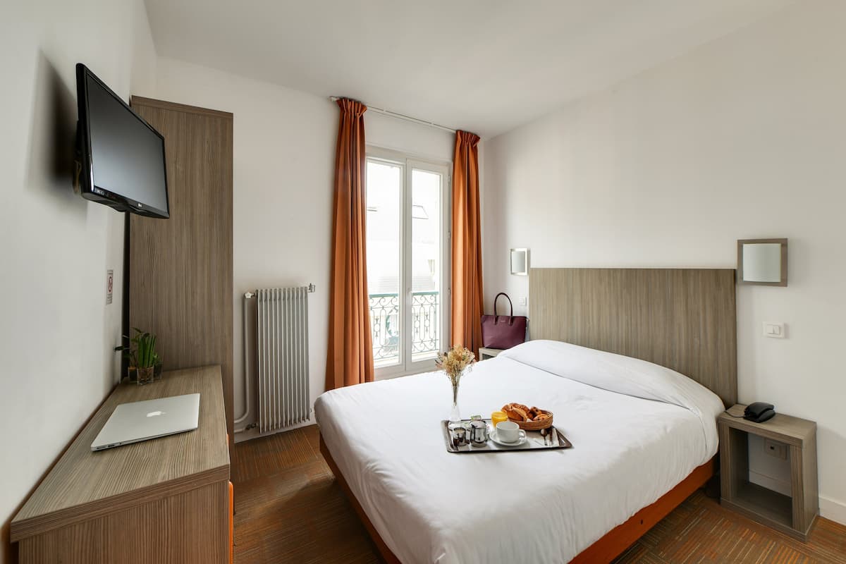 A hotel room with a bed, a desk with a laptop, a TV mounted on the wall, a radiator, and a window with curtains.
