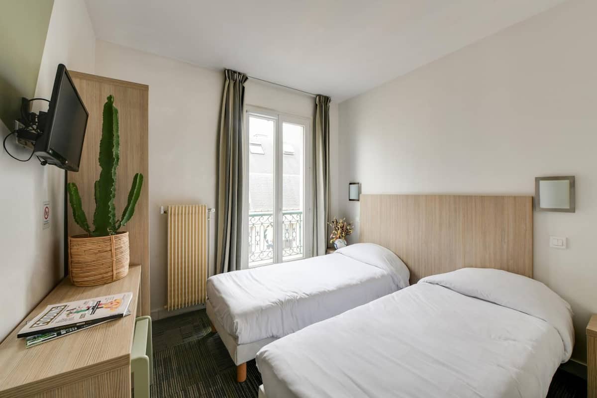 A hotel room with two single beds, a desk with a computer monitor, a potted plant, and a window with curtains.